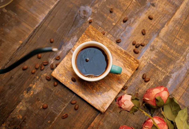 Cup of coffee on wood, surrounded by beans and roses